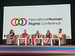 Scène d'une conférence sur les droits humains, avec cinq intervenants assis devant une diapositive