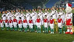 Sur un terrain de football, onze joueurs chantent leur hymne national, la main sur le cœur devant onze enfants.
