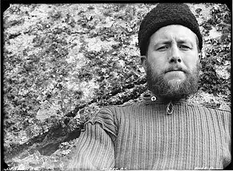 Photo noir et blanc, portrait en buste d'un homme portant la barbe et un gros bonnet sombre, devant une roche parsemée de lichens.