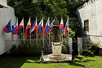 Statue jeune femme debout, vêtu d'une longue robe. Porte un drapeau plié dans ses mains, paumes tournées vers le ciel.