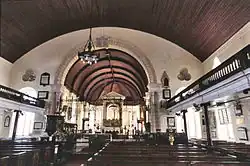 Photographie de l'intérieur d'une église : plafond voûté en bois avec poutres, murs blancs, de nombreuses rangées de bancs en bois sombre et poli avec une allée centrale menant à l'autel, balcons de chaque côté équipés de garde-corps et soutenus par des pilliers, deux grandes fenêtres voutées à l'arrière, lustre au plafond, autel en arrière centre comprenant un retable en or.
