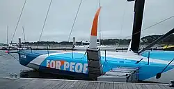 Vue latérale de l'avant d'un voilier bleu et blanc amarré au ponton. Le pont est bombé. Le bouchain est très marqué.