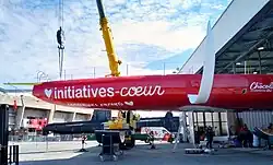 Profil bâbord de l'avant d'un voilier rouge à demi sorti de son hangar et soutenu par une grue. Le foil blanc bâbord est déjà en place.