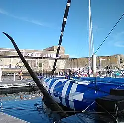 Vue de trois quarts arrière de l'avant d'un voilier bleu amarré au ponton. En premier plan, le foil bâbord.