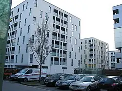 Vue de quelques bâtiments blancs identiques et carrés, ornés de nombreuses fenêtres rectangulaires, et d'un parking à l'avant-plan.