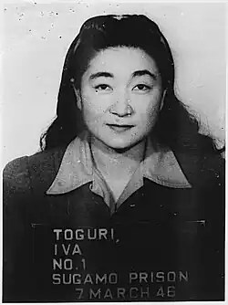 Photo d'une femme d'âge mur aux longs cheveux fixant le photographe en souriant. Le cadrage est typique des photos d'identification. Son nom et un numéro sont écrits sur une ardoise qu'elle porte devant elle.