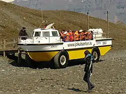 "Véhicule jaune et blanc a l’arrêt au sec devant un ponton, avec une vingtaine de personnes à bord portant des gilets de sécurité, dans un paysage minéral"