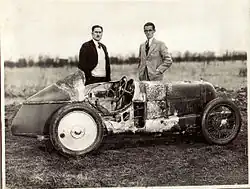Photo de deux hommes se tenant de pied derrière une voiture à la carrosserie chiffonée.