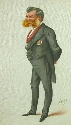 Portrait d'un homme en costume, avec une large barbe rousse et les mains derrière le dos.