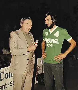 Deux hommes ebout, de face. Celui de gauche tend un micro vers celui-ci de droite, qui est barbu.