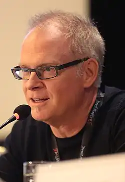 Photographie en couleurs d'un homme portant un pull noir et des lunettes, parlant devant un micro.