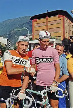 Photographie de deux cyclistes se donnant une poignée de main, l'un en maillot orange, l'autre en maillot rose.
