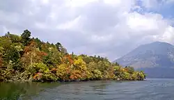 Photo couleur d'une pointe montagneuse et boisée, avancée dans une étendue d'eau (de gauche à droite). Un volcan (à droite) sous un ciel nuageux, en arrière-plan.