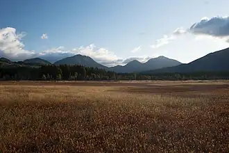 Photo couleur d'une pleine herbeuse aux couleurs automnales, avec, en arrière-plan, une chaîne de montagnes boisées, sous un ciel bleu nuageux.
