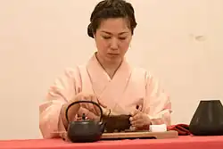 Une femme vêtue d’un kimono rose pâle. Devant elle, une théière et un plateau.