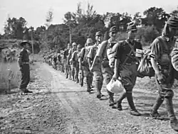 Photographie en noir et blanc de plusieurs militaires japonais marchant les uns derrière les autres sur un chemin en terre et sous la surveillance d'un militaire malaisien.