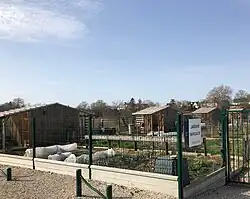 Photographie en couleurs d'un jardin, ceinturé d'un grillage, dans lequel se trouvent un cabanon et des tunnels de serres.