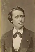 photo à couleur sépia : un homme aux yeux clairs, au visage allongé, aux traits fins, portant costume et nœud papillon, regarde au loin sur sa droite.