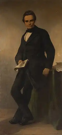 Joseph Lebeau, élégamment vêtu, pose de trois quarts. Il tient un papier de la main droite et est appuyé contre un meuble.