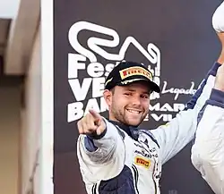 Photographie d'un homme debout, de face, qui pointe vers la caméra avec un sourire