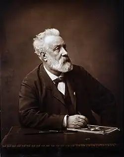 Photo noir et blanc de Jules Verne, un homme barbu aux cheveux blancs assis devant une table, le crayon à la main.