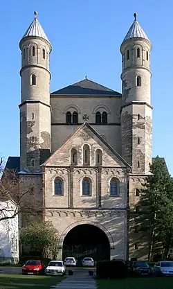 Façade d'église à vaste porche surmonté d'un large et haut fronton triangulaire, devant une massive tour carrée flanquée de deux tours plus élevées, étroites et cylindrique