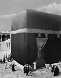 La Kaaba en 1910. On voit bien le seuil qui surélève la porte d'entrée, et on distingue, dans l'angle, la Pierre noire derrière le groupe d'hommes.
