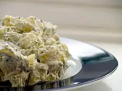 Vue partielle d’une assiette à bord argenté et fond blanc où se trouve un gros tas de morceaux de pomme de terre à la mayonnaise et au persil haché.