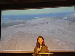 Photographie d'une femme parlant au micro devant des images de pollution.