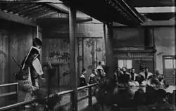 Photo noir et blanc de 1943. Au premier plan à gauche, l'acteur s'avance sur le pont ; à droite, les spectateurs vus de dos. Au fond à gauche, la paroi est ornée de bambous stylisés, à droite, musiciens accroupis sur la scène.