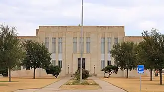 Vue partielle de la façade d'un bâtiment de deux étages. Des arbres et un mat sont présents au premier plan.
