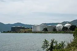 Photographie d'une usine pétrolière et de ses nombreuses citernes. L'ensemble est entouré par la mer au premier plan et par les montagnes à l'arrière-plan.