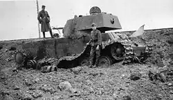 Photo en noir et blanc d'un char détruit. Un soldat se tient par terre, à côté de la chenille détruite, un autre est debout sur le chassis du tank, devant sa tourelle. Le char est entouré de débris.