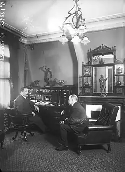 Photographie en noir et blanc d'hommes en costumes assis sur des fauteuils, discutant à un bureau.