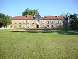 Un grand terrain vert avec à l'arrière-plan un long bâtiment à deux étages, de pierre ocre au toit de tuiles.