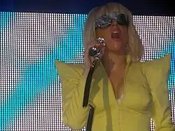 Une femme vêtue d'un veston jaune est visible. Ses cheveux sont en forme de bob et elle porte une paire de lunettes. Elle tient un microphone dans sa main droite qui va jusqu'à sa bouche. Le fond de l'image montre un écran avec des motifs indigos et blancs.