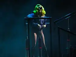 Photographie de Lady Gaga debout sur une plateforme, vêtue d'une veste mauve et d'une paire de lunettes de la même couleur.