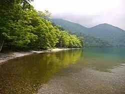Photo couleur d'un lac à l'eau transparente. Au second plan, s'étend le littoral boisé du lac, sous un ciel chargé de nuages.