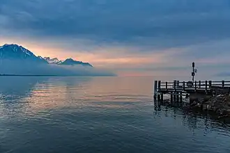 Photo du lac, sur la droite un ponton, à l'arrière plan les alpes.