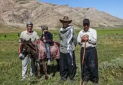 Un enfant avec un bâton sculpté assis sur un âne entouré de trois adultes d'âges variés se tenant debout, l'un appuyé sur un bâton, dans un champ en altitude.