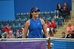 Une joueuse marche sur un terrain de tennis vers le filet, souriante. Au fonds, des gradins parsemés de spectateurs.