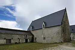 Photographie en couleurs et en trois quarts d'un bâtiment à toiture pentue recouverte d'ardoises munie de fenêtres à meneaux, une seconde construction formant un angle droit visible à gauche.