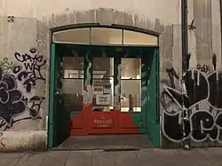  une porte qui donne sur une rue, avec 3 battans vitrés. Il y a des grafiti colorés sur les murs de chaque côté. Dans la partie inférieure de la porte centrale, on peut lire Le refuge Genève