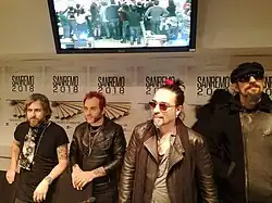 Au premier plan, quatre personnes en ligne horizontale, derrière eux, cinq panneaux rectangulaires avec des textes et des chiffres, au-dessus, une fine corniche surmontée centralement d'un grand écran tronqué avec une image.