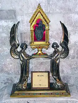 Deux anges ailés tiennent une boite présentant des ouvertures vitrées sur les côtés et contenant une main momifiée.