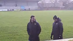 Photo de deux hommes debout devant un terrain de football. Au fond se trouve une tribune de stade.