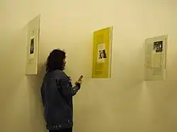 Une femme avec un smartphone devant 3 panneaux de verre perpendiculaires à un mur.