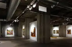 Photographie en couleurs de la salle principale vue d'un angle, des piliers de béton qui la parcourent et de quelques œuvres.