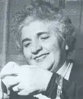 Image en noir et blanc d'un visage de femme, cheveux frisés blancs regardant avec un sourire un objet tenu dans sa main.