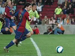 Photographie couleur. Au premier plan, Lionel Messi court pour le FC Barcelone sur un terrain. Il dispute un ballon à la lisière de la surface de réparation avec un adversaire revêtant les couleurs du Getafe FC, situé au second plan. À l’arrière plan, une tribune accueillant un public clairsemé.